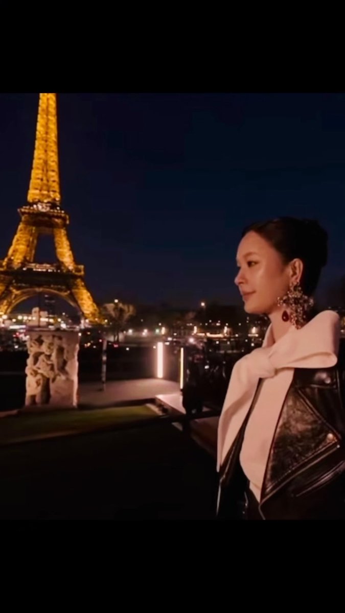 A woman with dark hair, wearing a light scarf and dark leather jacket with large earrings, stands with the illuminated Eiffel Tower in the background at night.