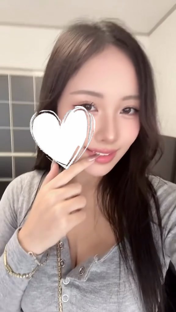 A young woman with long dark hair wearing a grey top smiles and points at her face, which is partially obscured by a white heart graphic.