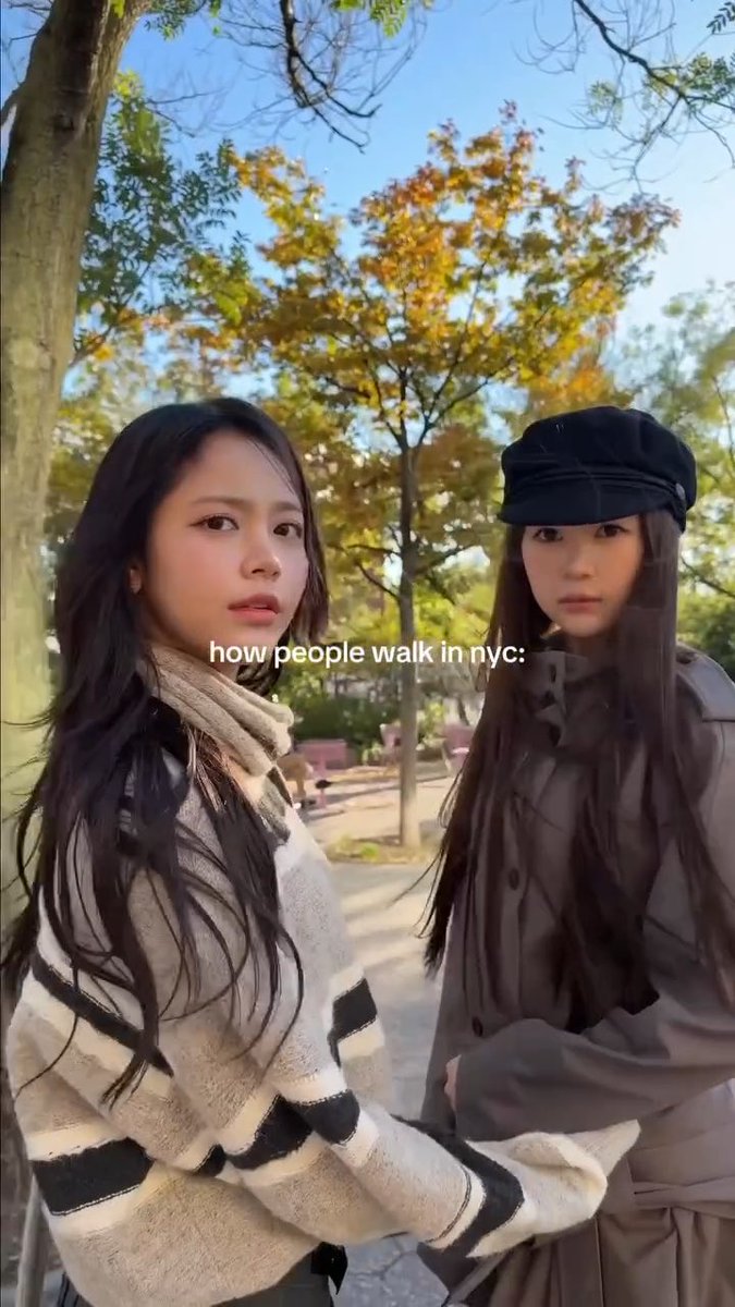 TikTok-style video still with text 'how people walk in nyc:' showing two young women in a park wearing coats, with autumn trees visible in the background.