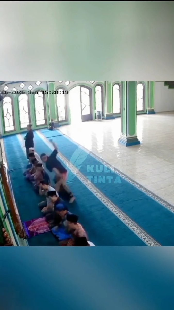 A security camera screenshot showing people engaged in what appears to be a physical altercation or takedown on a green carpet inside a mosque.