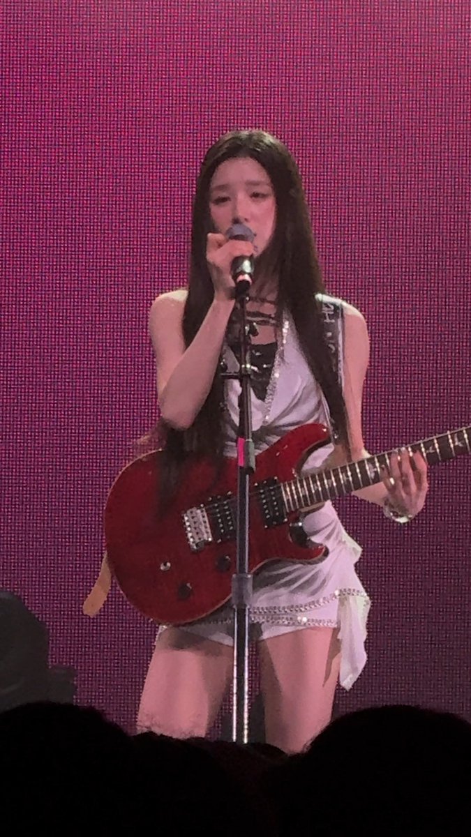 A young woman performing on stage with a red electric guitar, wearing a light-colored sleeveless outfit against a pink/magenta background.