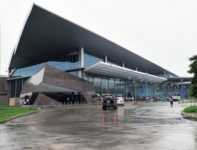 The exterior of a large modern building with an angular, wing-like roof and glass facade is shown on a rainy day with cars and pedestrians outside.