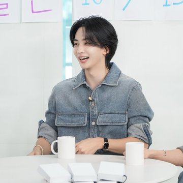 A smiling young man with dark hair in a denim jacket sitting at a table with white mugs during what appears to be a talk show or interview setting.