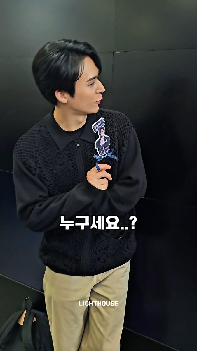 A young man in a black textured vest and khaki pants holds an acrylic fan photo, with the text '누구세요..?' (Who are you..?) and 'LIGHTHOUSE' watermark overlaid.