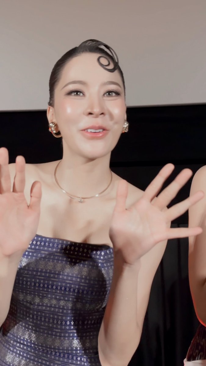 A woman with sleek hair styled in a curled front piece waves at the camera, wearing a dark patterned strapless top and silver hoop earrings.