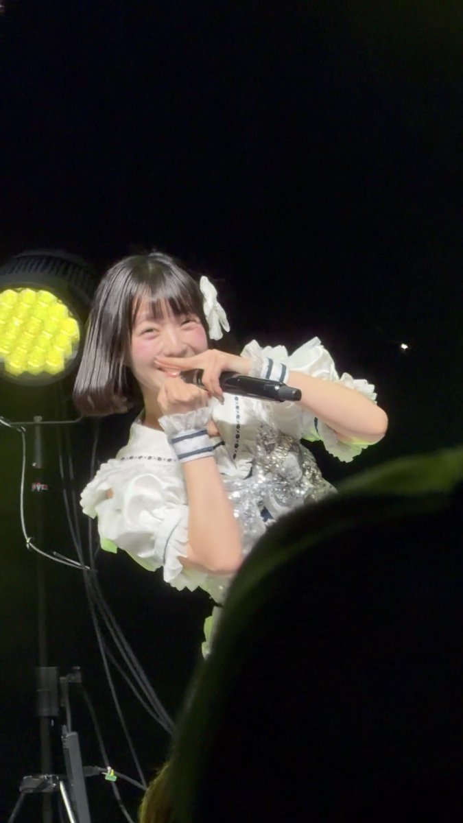 A young Japanese performer with short dark hair and a white bow, wearing a ruffled white idol outfit, smiling while holding a microphone on a dark stage with stage lighting.
