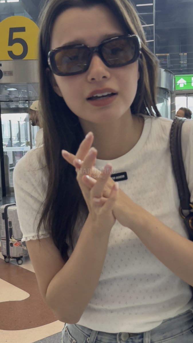 A young woman wearing large black sunglasses and a white textured top, posing at what appears to be an airport with a yellow "5" sign visible in the background and a watermark reading "HARU HARU haruharu05_".