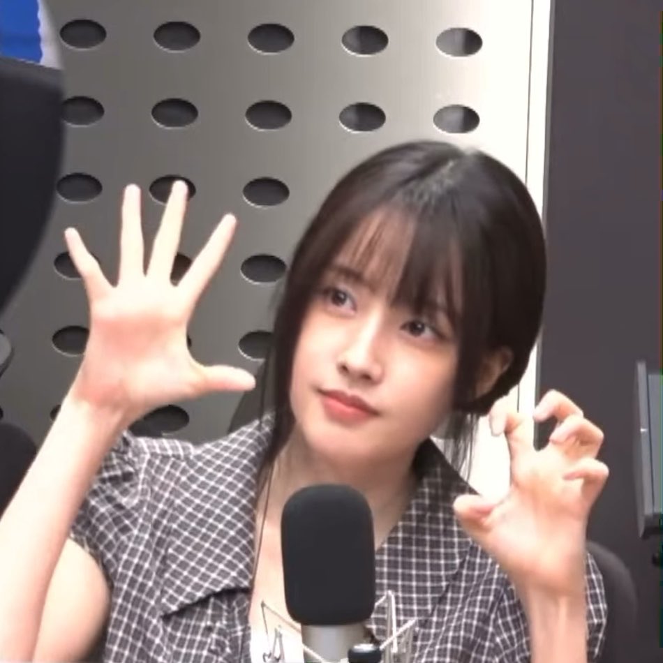 A young woman with dark hair and bangs sits in front of a microphone in a studio with soundproofing, holding her hands up playfully.