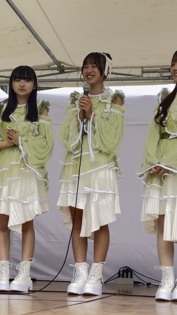 Three young women in matching green and white frilly dresses and white boots standing under a tent, with the middle one holding a microphone.