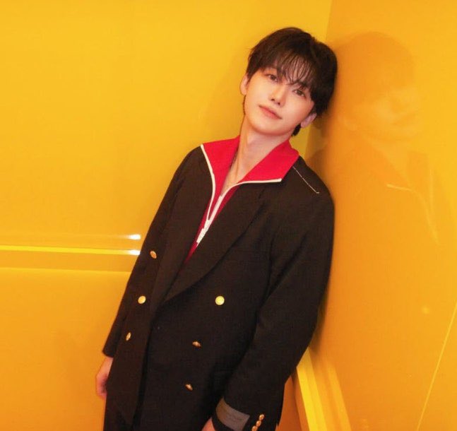 A young man with dark hair leans against a bright yellow wall, wearing a black double-breasted blazer with gold buttons over a red and white collared shirt.