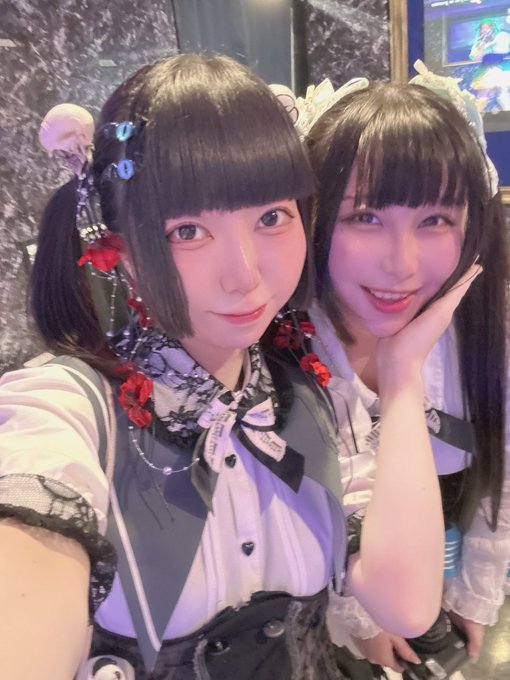 Two young women pose together, one with short black hair in a Gothic-style outfit with red roses and the other with long dark hair in a maid-style headband.