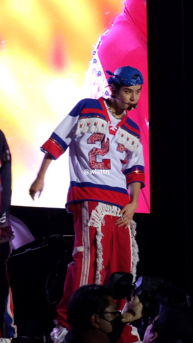 A male performer on stage wearing a red, white, and blue hockey jersey with fringe, red pants, and a blue headpiece with a headset microphone, credited to @wisttii.