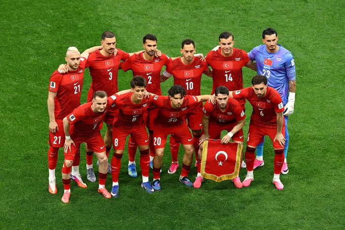 The Turkish national football team poses for a pre-match team photo on the pitch, with one player proudly holding a small Turkish flag. The red kits display visible jersey numbers including 3, 2, 10, 14, 21, 8, 20, and 6, alongside the team's goalkeeper in a blue #23 jersey. The caption "Our Cacıklar" is a playful Turkish slang reference, using the word for a traditional yogurt dish to affectionately refer to the squad.