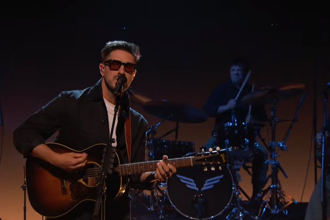 A musician wearing sunglasses and a black jacket plays an acoustic guitar and sings into a microphone during a live performance, with a drummer visible in the background. This image captures Mumford & Sons' appearance on "Jimmy Kimmel Live," where they performed their song "Here."