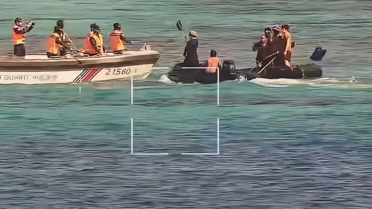 An aerial view showing two boats in the water—one a white Coast Guard vessel marked '21560 1' and the other a black inflatable boat—along with a cameraman filming the scene, with a white camera viewfinder overlay visible.