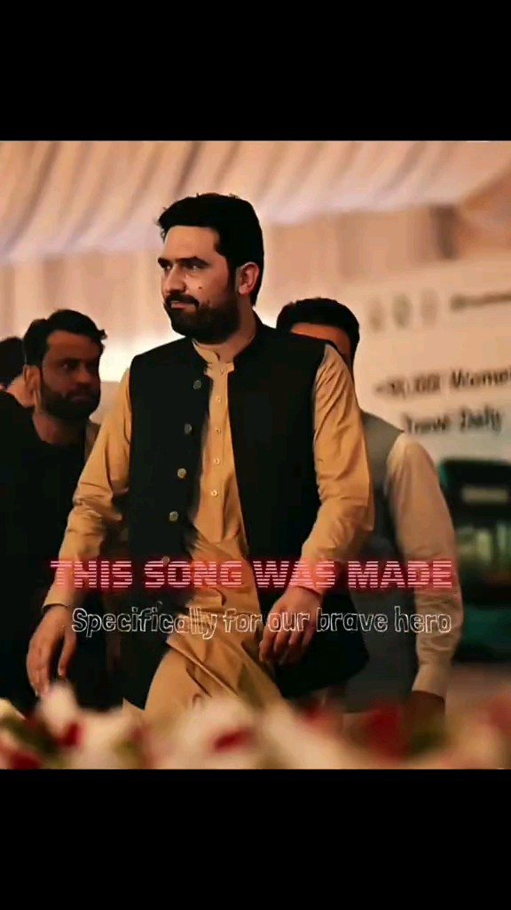 A man in a beige shirt and black vest at what appears to be a memorial gathering, with overlaid text reading 'THIS SONG WAS MADE' and 'Specifically for our brave hero'.