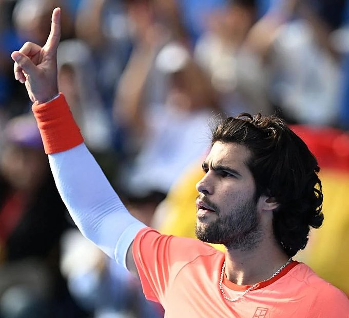A dark-haired, bearded tennis player wearing a coral Nike shirt and wristband points skyward in celebration, likely after a key victory. The image accompanies a post highlighting Carlos Alcaraz's elite 81.84% win rate, ranking him fifth among the most effective male tennis players of the Open Era.