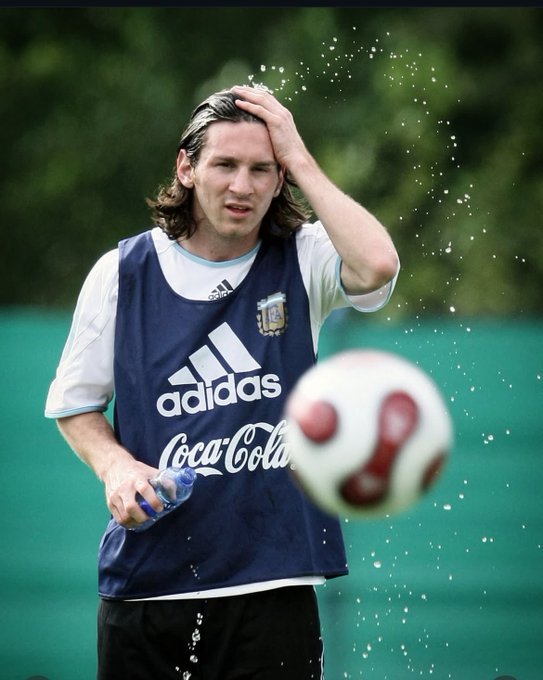 A long-haired soccer player in an Argentina Adidas training bib sprays water on his head while heading a ball during practice.