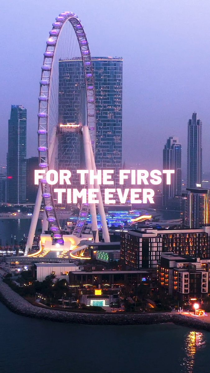 An aerial nighttime view of a large illuminated Ferris wheel in a city skyline, with text overlay reading 'FOR THE FIRST TIME EVER'.