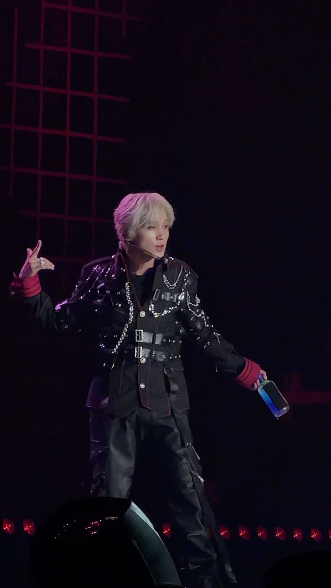 A performer with pink hair, wearing a black studded jacket with chains, stands on a stage with a dark background and red grid lights, holding a blue phone-like object.