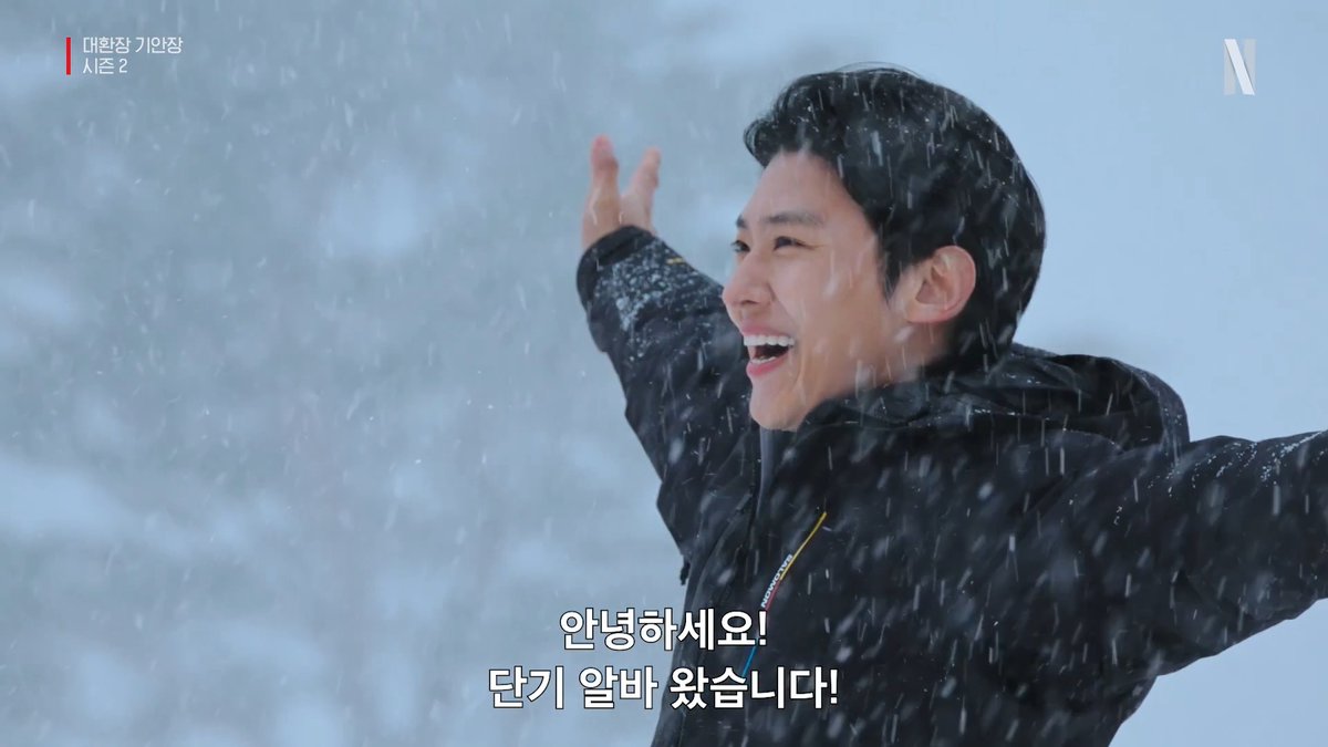 A man in a black winter jacket stands joyfully with his arms outstretched in heavy falling snow, with Korean subtitles reading "안녕하세요! 단기 알바 왔습니다!" (Hello! I'm here for the short-term part-time job!), capturing a scene from the Netflix series "대환장 기안장" (Grand Insanity Proposal) Season 2.