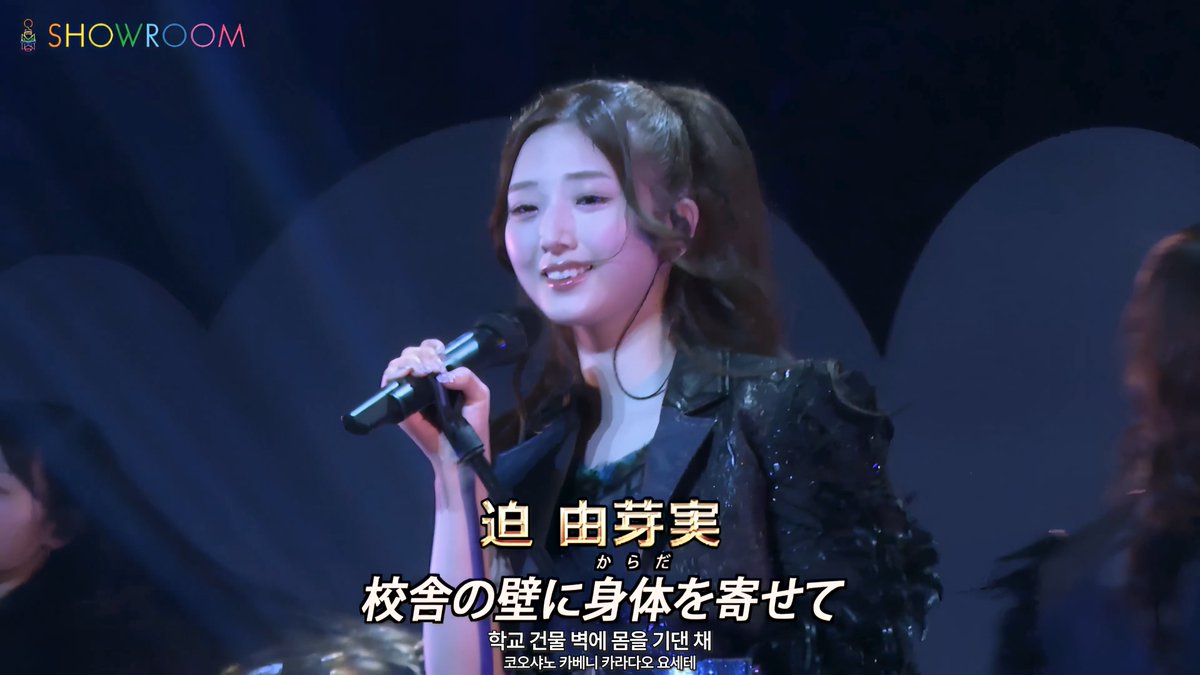 A female performer singing on a stage lit in blue tones, identified by yellow Japanese text as 迫 由芽実 (Sako Yumei) from SHOWROOM, with Japanese and Korean subtitles displayed.