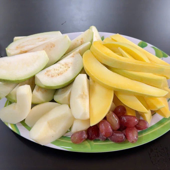 A plate filled with freshly cut fruit, including green guava, yellow mango slices, white melon or apple pieces, and red grapes.