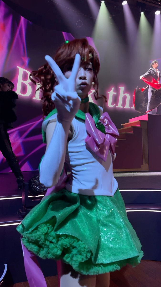Cosplayer dressed as Sailor Jupiter from Sailor Moon on stage, making a peace sign while wearing the green and white sailor fuku costume with a headset.