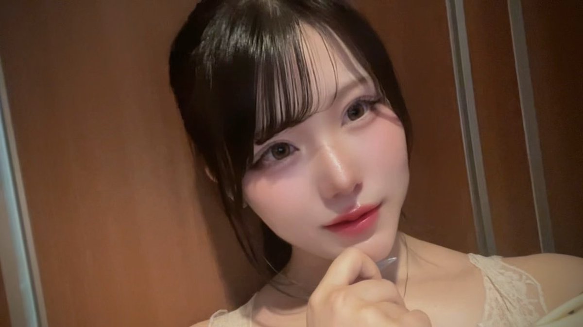 A close-up selfie of a young woman with dark hair and bangs wearing a cream lace top, posing with her hand near her chin.
