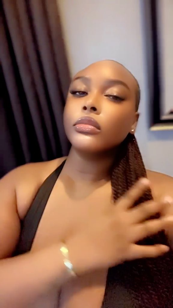 A woman with a shaved head and long braided ponytail poses confidently in a black halter top, wearing gold jewelry and glossy makeup, complementing the caption's message of unwavering compliance and devotion.