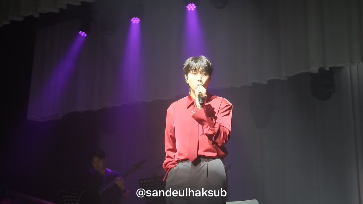 A young man in a red shirt and gray trousers singing into a microphone on a stage with purple lighting, with the watermark '@sandeulhaksub' visible at the bottom.
