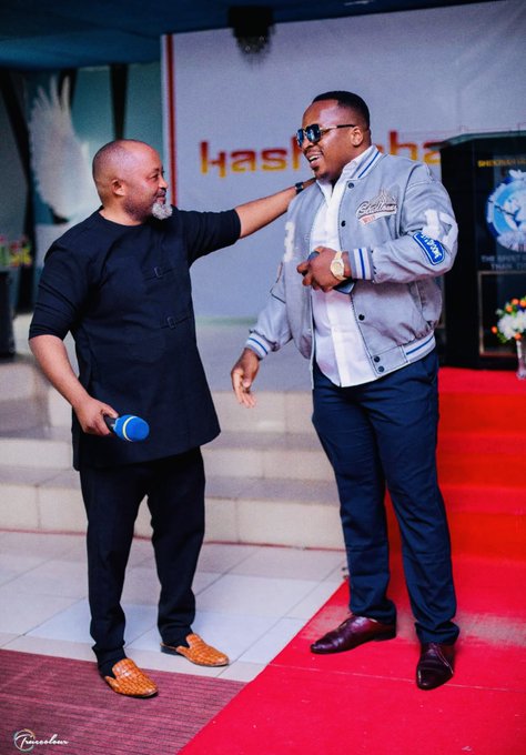 Two men laughing together at an event; the man on the left wears a black tunic and holds a blue microphone, while the taller man on the right wears a gray letterman jacket, sunglasses, and dark pants.