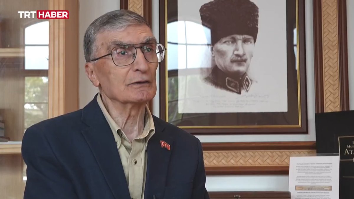 Nobel Prize-winning Turkish scientist Prof. Dr. Aziz Sancar speaks during a TRT Haber interview, seated in front of a framed portrait of Mustafa Kemal Atatürk. The Turkish flag pin on his lapel complements his message urging Turkish scientists to study abroad but return to serve their homeland.