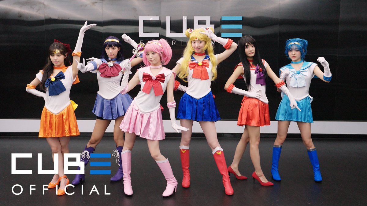 Six women in colorful Sailor Moon cosplay outfits pose together in front of a "CUBE ENTERTAINMENT" backdrop, with a "CUBE OFFICIAL" watermark in the lower left corner.