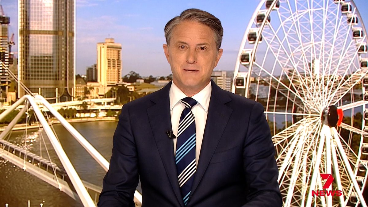 A male news anchor in a dark suit and striped tie delivers a broadcast for "7 NEWS" against a city skyline featuring a large Ferris wheel and bridge.