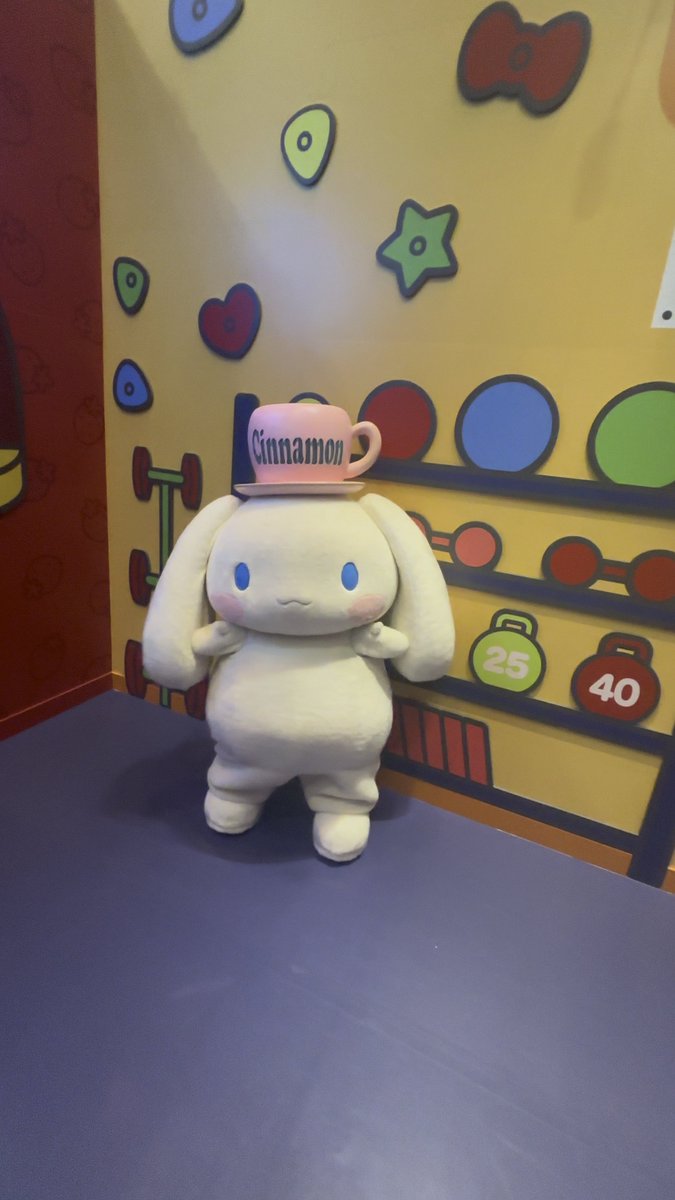 A white plush rabbit character 'Cinnamoroll' mascot wearing a pink 'Cinnamon' cup on its head, standing in a colorful room with playful wall decorations including stars and numbers.