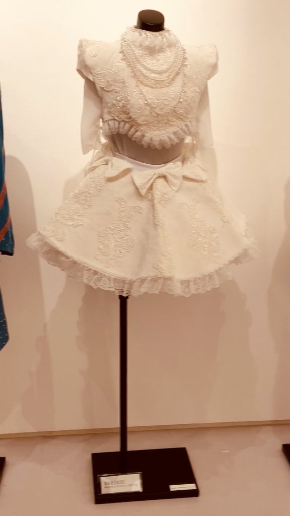 A white lace two-piece outfit consisting of a crop top and skirt with a bow is displayed on a mannequin in what appears to be a gallery setting.