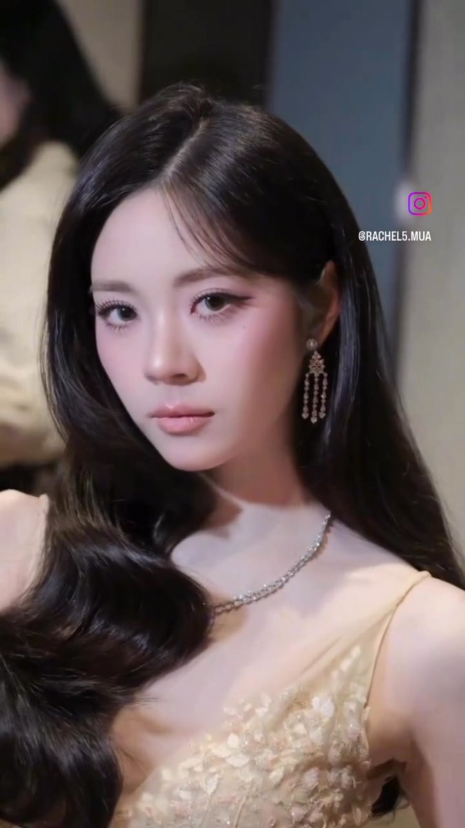 A close-up portrait of a young woman with long dark hair wearing a yellow dress, a delicate necklace, and dangling earrings, with the watermark '@RACHEL5.MUA' visible.
