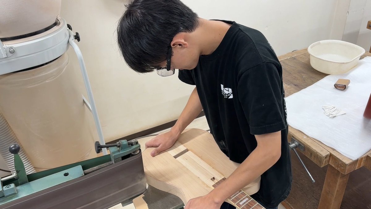 A young man wearing glasses and a black t-shirt is working on shaping a wooden guitar body using a sanding machine in a workshop.