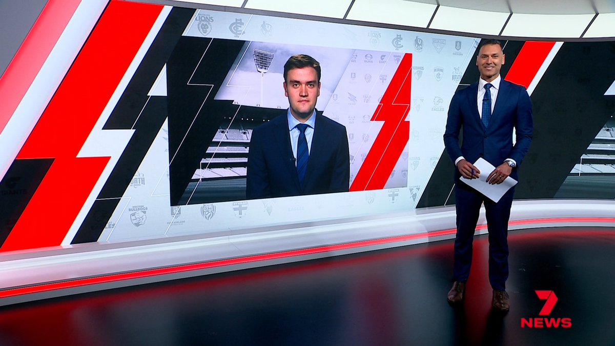 Two male news anchors in blue suits appear in a modern Channel 7 News studio with red and black geometric design elements, featuring an AFL team logo backdrop on their screens.