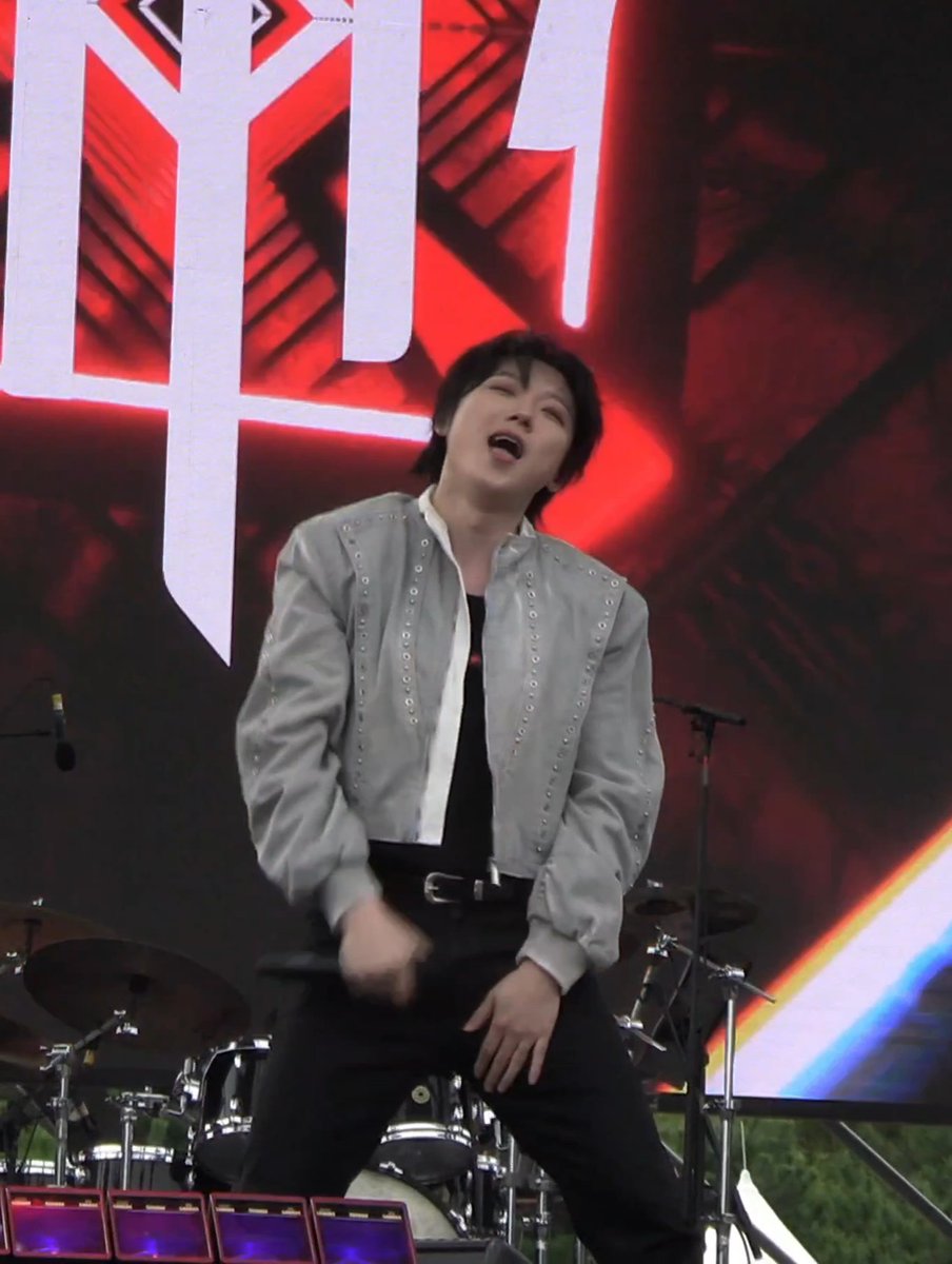 Image 8: "A man in a gray jacket with studs performs on stage with his eyes closed and mouth open, in front of a large red and white background."