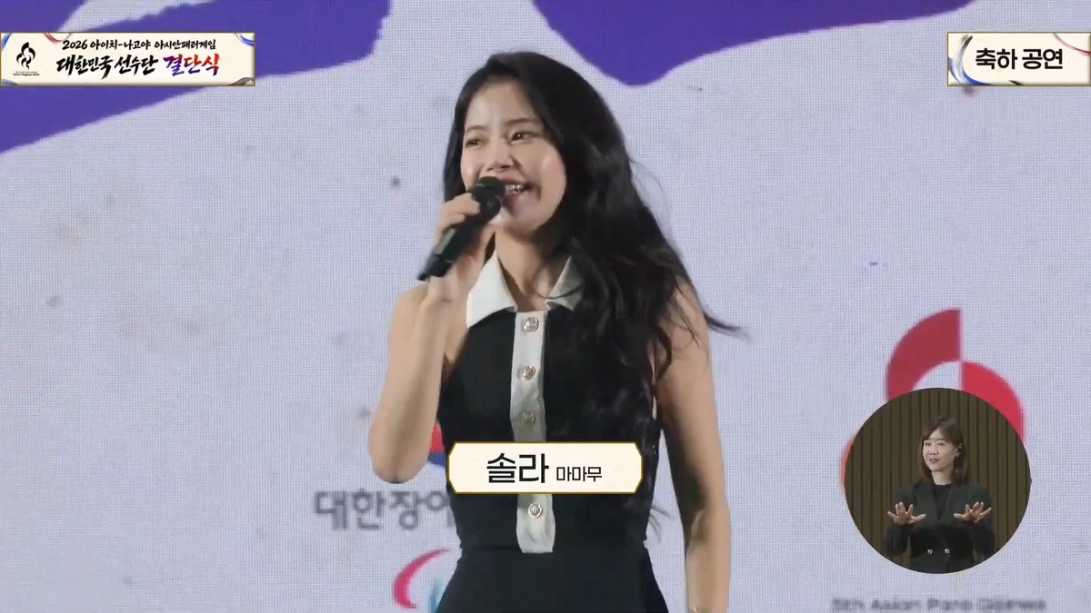 South Korean broadcast showing singer IU performing with a microphone, identified by a label reading '술라 아이유', alongside a sign language interpreter.
