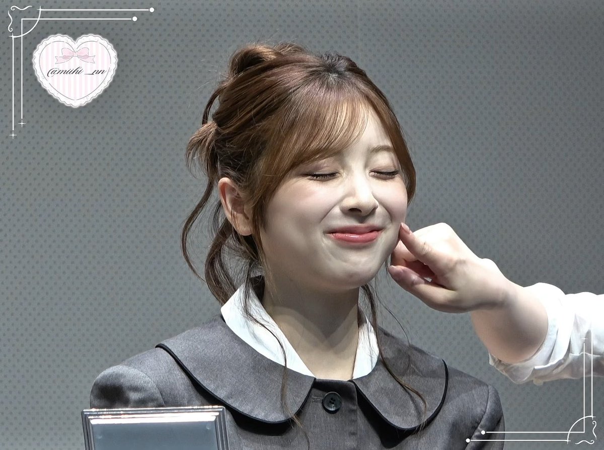 A smiling woman with her eyes closed and brown hair in an updo wears a gray outfit with a white collar, while another person's hand touches her cheek.