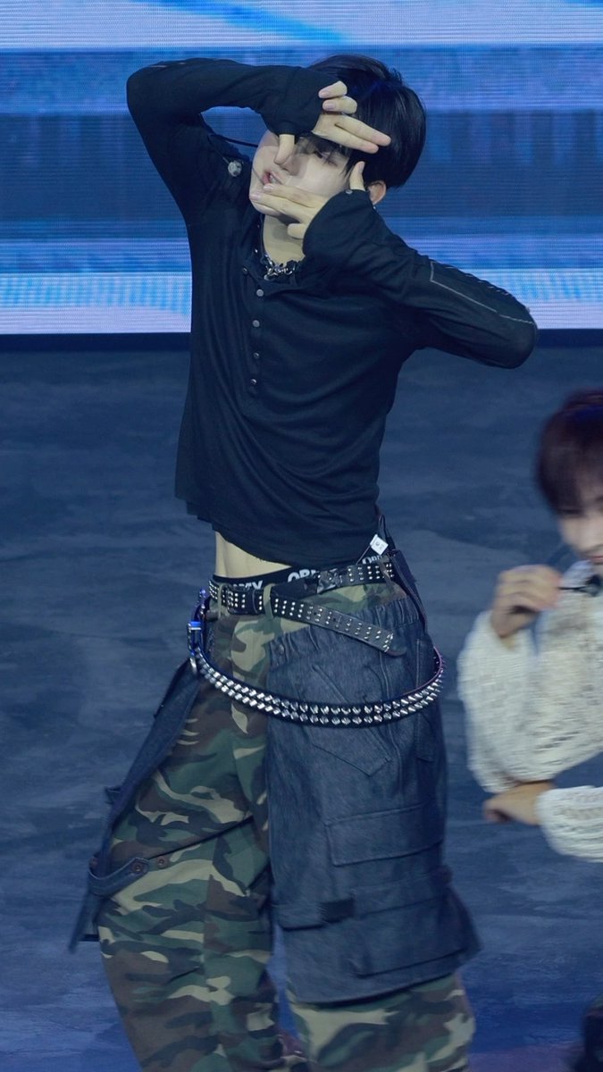 A performer on stage wearing a black sheer top, studded belts, and camouflage pants, posing with one hand over their face.