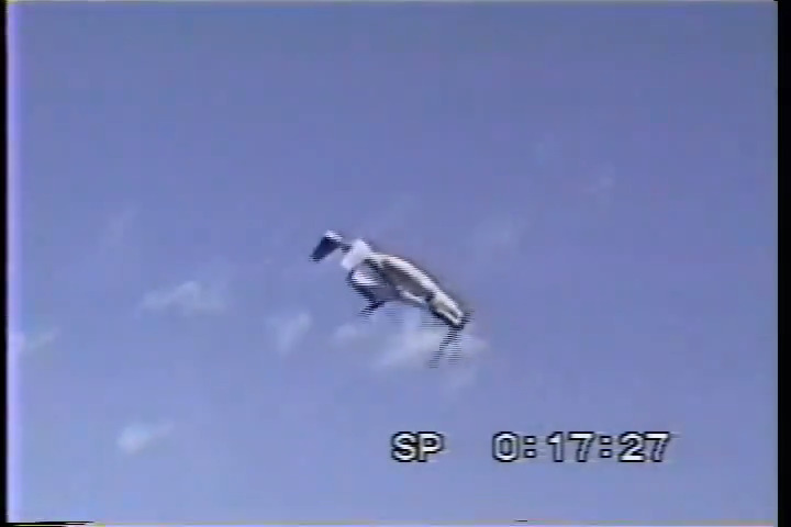 A V-22 Osprey tiltrotor aircraft is captured mid-aileron roll against a blue sky with scattered clouds, its fuselage inverted with wings and rotors visible. The bottom of the frame shows "SP 08:17:27" in a digital timestamp overlay, characteristic of camcorder home video footage from the late 1990s or early 2000s. The image directly illustrates the caption by showing the unusual and dramatic maneuver of a V-22 performing a full rotation in flight.