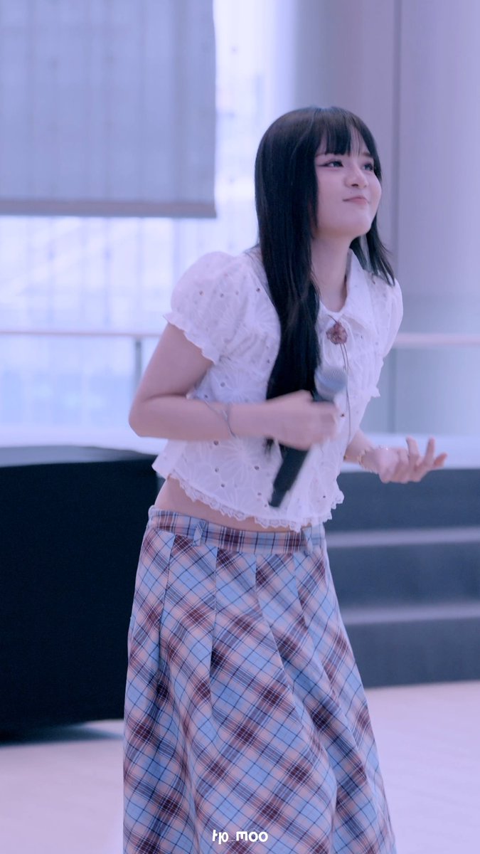 A young woman with long dark hair wearing a white eyelet crop top and plaid pants, holding a microphone and smiling while looking to the side, with a watermark reading 'ho_moo'.