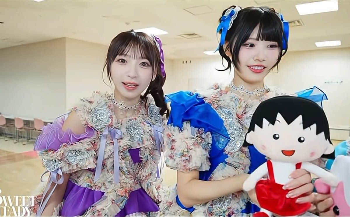 Two Japanese idols in elaborate pastel frilly costumes with ribbons, one in purple and one in blue, posing with a Chibi Maruko-chan doll, with a 'SWEET READY' watermark visible.