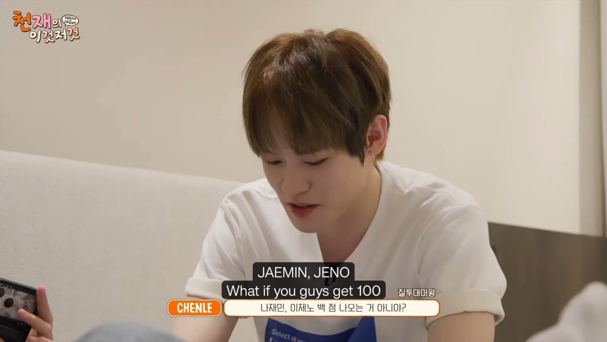 A young man in a white t-shirt sits on a couch looking at a phone, with subtitles showing Chenle asking Jaemin and Jeno 'What if you guys get 100' (절두대미왕 '나재민, 이제는 백 점 나오는 거 아니야?').