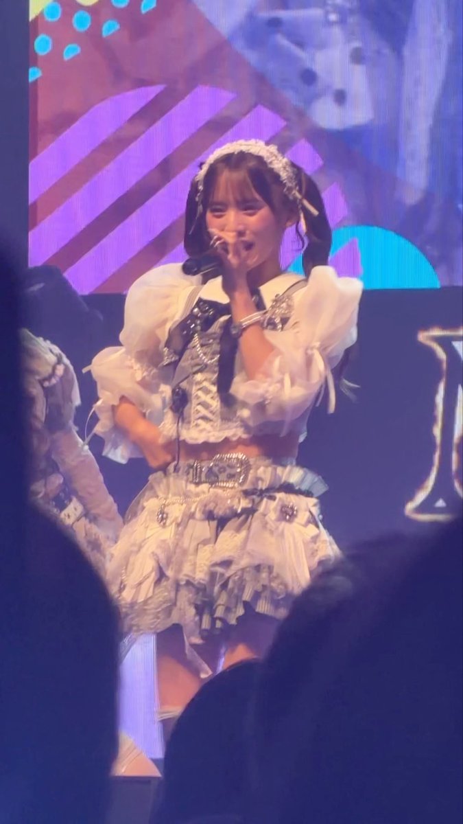 A performer in an elaborate white and silver frilly stage costume with pigtails sings into a microphone against a colorful patterned backdrop.