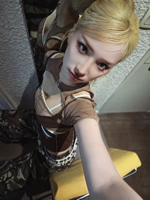 A selfie of a blonde person wearing a brown outfit with multiple cutouts and a studded belt, looking up at the camera from a high angle.